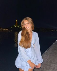 a beautiful young woman standing on top of a pier next to the ocean at night