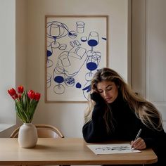 a woman sitting at a table writing in front of a vase with red tulips