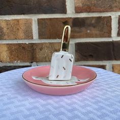 a white and pink plate with a gold handle on it sitting on a table next to a brick wall