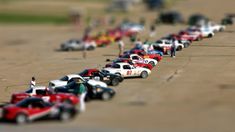 many toy cars are lined up in a row on the pavement, with people standing around them