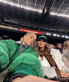 two women sitting next to each other in the stands at a baseball game, one wearing a green hat