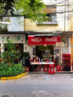 two people sitting at a table in front of a building with red chairs on the sidewalk