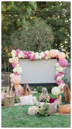 two women sitting on the grass watching a movie in front of a large screen with balloons