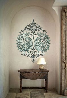 a room with a table and lamp next to a painting on the wall that has an ornate design