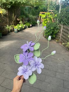 a hand is holding a crocheted purple flower in the middle of a walkway