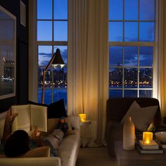 a person laying on a couch reading a book in front of two large windows at night