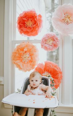 a baby sitting in a high chair with flowers hanging from the window behind it,