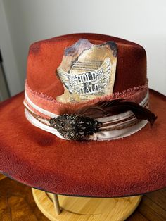 "The perfect burnt orange (rust) hat has been hand painted, and distressed. It's a smaller fedora and has a pink velvet band, wrapped in lace and gold threading. A vintage iron pin is attached. Tied with pink twine is a red feather and brown duck tail feathers.   SIZE: Fedora Hat is OSFA Wired brim measuring 3 3/4\" and a crown height of 4 3/4\". The Fedora Hat has a adjustable band with drawstring which allows for a perfect fit. Materials: This hat is made from a blend of 65% cotton and 35% pol Vintage Distressed Hats For Country Events, Vintage Distressed Hats For Rodeo, Distressed Brown Vintage Hat, Vintage Distressed Hat For Western-themed Events, Vintage Fedora For Country Events, Vintage Felt Hat With Short Brim For Festivals, Vintage Handmade Hat Band For Western-themed Events, Vintage Handmade Hats For Western-themed Events, Vintage Handmade Hat For Western-themed Events