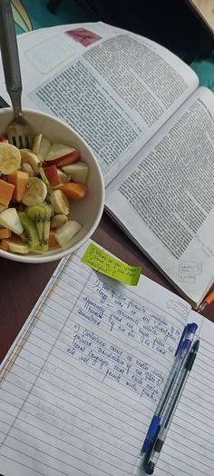 a bowl of fruit sitting on top of a table next to an open book and pen