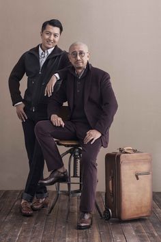 two men sitting on top of a chair next to a piece of luggage in front of them