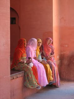 three women sitting on a ledge with their heads turned to the side, wearing brightly colored clothing