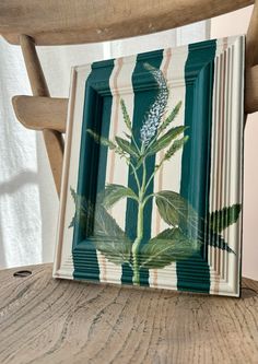 a painting on a wooden table with a chair in front of it and a plant painted on the frame