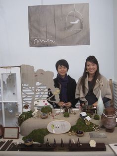 two women sitting at a table covered in miniature trees and plants, surrounded by other items