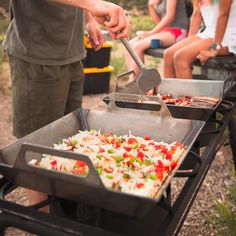 some people are sitting in the grass and one is grilling food on an outdoor grill