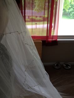 a white wedding dress hanging in front of a window with red curtains and shoes on the floor