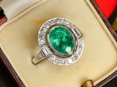 Grassy green and undertones of blue punctuate this vision of an Art Deco inspired diamond ring. A large oval, faceted emerald of 2.56 carats exudes saturated hues and at its depth, bright highlights. It is bezel set, then after an airy opening, encircled by brilliant diamonds.  Not only are single-cut diamonds utilized, but two rectangular baguettes and two tapered baguettes touch off each shoulder.  A handmade basket weave under-gallery allows light to penetrate in order to bring out the best of the stones.  We dare you to compare prices for anything similar for this ring's quality, size, and attributes! Details: One oval faceted natural emerald Weight of 2.56 carats Twenty diamonds Total weight of .84 carats Color G-H for baguettes Clarity VS Color H-I for single cuts Clarity VS2-SI1, tw Oval Hallmarked Emerald Ring In Platinum, Hallmarked Oval Emerald Ring In Platinum, Gia Certified Oval Emerald Ring In Art Deco Style, Art Deco Oval Emerald Ring With Brilliant Cut, Green Oval Emerald Ring In Platinum, Platinum Oval Emerald Ring With 17 Jewels, Oval Platinum Emerald Ring With 17 Jewels, White Gold Oval Emerald Ring Art Deco Style, Art Deco Oval Jewelry With Halo Setting