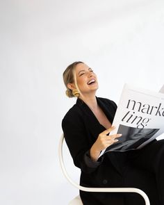 a woman sitting in a chair holding a magazine and smiling at the camera while laughing