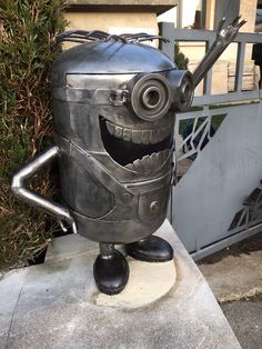 a metal robot statue sitting on top of a cement slab next to a gate and bushes