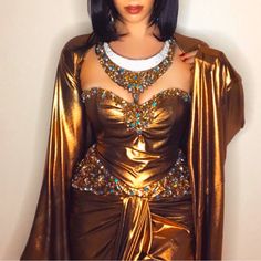 a woman dressed in gold and wearing a costume with jewels on her neck, standing against a wall