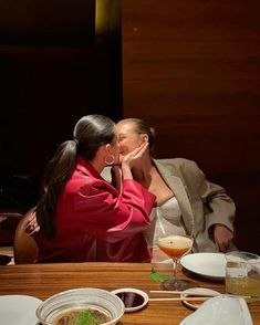 two women sitting at a table with food and drinks in front of them, one is kissing the other's face