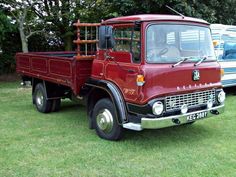 an old red truck parked on top of a lush green field next to other trucks