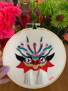 a person holding up a hand embroidered artwork on a wooden table with flowers in the background