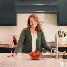 a woman standing in a kitchen next to a bowl of fruit and a glass of wine