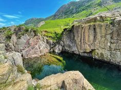 a body of water surrounded by large rocks and green grass on the side of a mountain