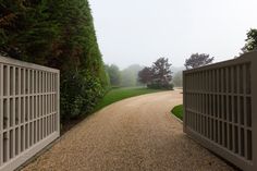 a gravel road between two gates leading to a lush green field with trees in the background