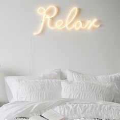 a bed with white sheets, pillows and a neon sign above it that reads relax