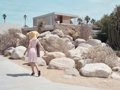 a woman wearing a hat walking down a sidewalk next to large rocks and cactus trees