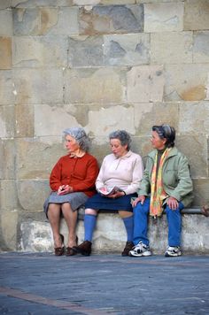 three older women sitting on a bench in front of a stone wall and looking at something