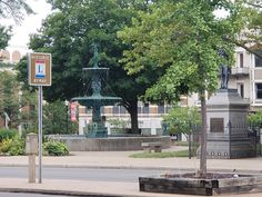 a street corner with a fountain in the middle and trees on both sides, surrounded by buildings