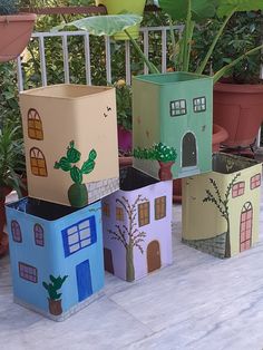 four cardboard boxes with houses painted on them sitting in front of some potted plants