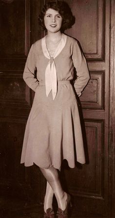 an old photo of a woman standing in front of a door wearing a dress and tie