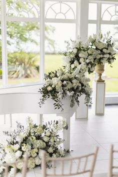 white flowers and greenery are arranged on the back of a piano in front of large windows