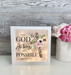 a white vase with flowers and a cross on it next to a sign that says, with god all things are possible