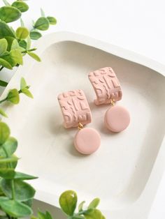 two pink and gold earrings sitting on top of a white tray next to green plants