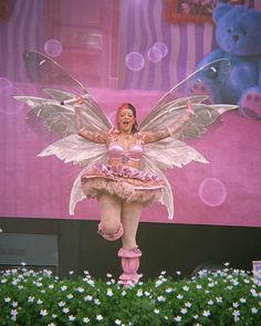 a woman dressed as a fairy standing on top of a flower bed in front of a giant screen