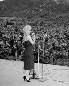 a woman standing at a microphone in front of a large group of people with mountains in the background