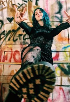 a woman with blue hair posing in front of graffiti covered wall and holding her hands up