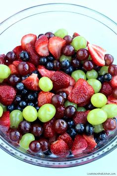 a glass bowl filled with grapes, strawberries and other fruit on top of a white table