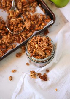 an apple and granola mix in a glass jar next to a baking pan with spoons