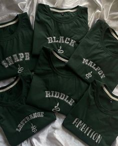 three green shirts with white letters on them are laying on a bed and one is black