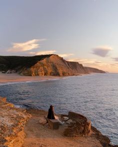 a woman sitting on the edge of a cliff by the ocean at sunset or sunrise