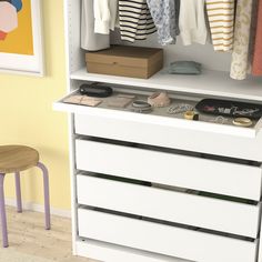 a white cabinet with drawers and clothes hanging on the wall next to a small stool