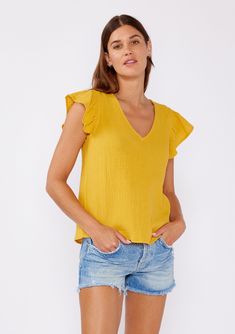 [Color: Mustard] An image of a brunette model wearing a mustard yellow top crafted from cotton gauze. With short flutter sleeves and v neckline. A comfortable top for the summer to fall season that can be paired with denim shorts and pants. Spring Short Sleeve Cotton Gauze Tops, Spring Cotton Gauze Short Sleeve Tops, Chic Summer Tops With Flutter Sleeves, Casual Butterfly Sleeve Blouse For Summer, Cotton Butterfly Sleeve Top For Summer, Spring Cotton Gauze Tops With Relaxed Fit, Summer Cotton Top With Butterfly Sleeves, Summer Cotton Tops With Butterfly Sleeves, Spring Relaxed Fit Cotton Gauze Tops