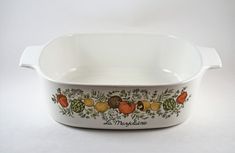 a white casserole dish with fruits and vegetables painted on the side, in front of a white background