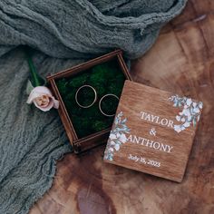 a wooden box with two wedding rings in it next to a flower and a blanket