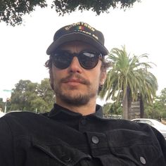 a man wearing sunglasses and a hat standing in front of a palm tree with his arms crossed